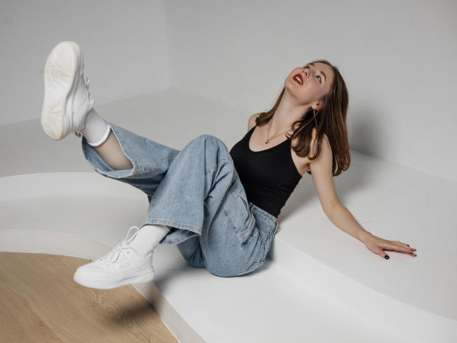MarieLoving is in a black tank top and loose, light blue jeans. She's sitting on a white, curved surface with her back on the floor, legs up and feet in white sneakers. Her head is tilted back, mouth open, and she has a red lipstick. She has light brown hair, white socks, and black nail polish. The image has a casual, contemporary style with a focus on relaxed, modern fashion. The composition is simple, highlighting MarieLoving's relaxed pose and stylish outfit.