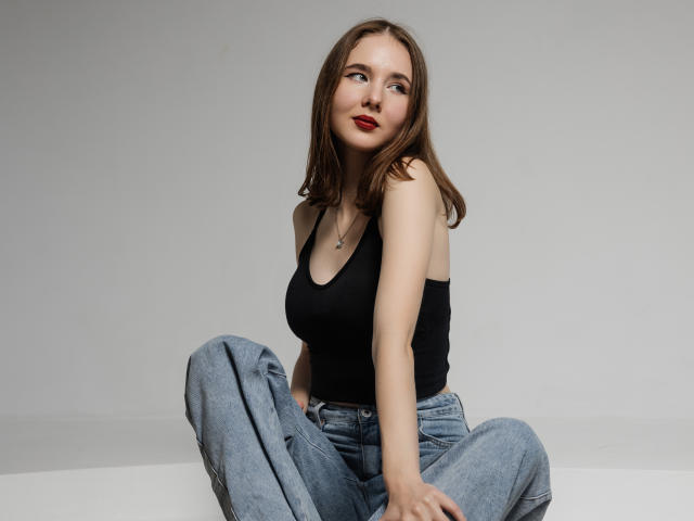 MarieLoving with small boobs, sitting cross-legged, wearing a black tank top and blue jeans, has red lipstick, and a necklace, looking to the side with a slight smile.