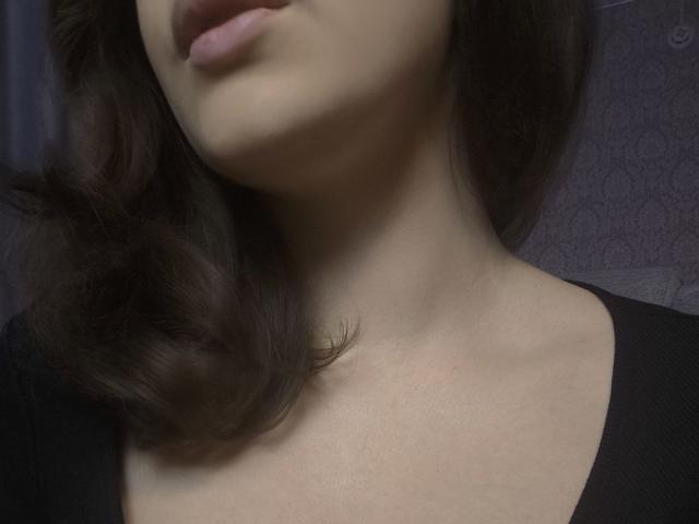 Dreia's neck and upper chest with wavy brown hair, pinkish lips, and fair skin, wearing a black top. Boobs partially visible, collarbone highlighted. Soft lighting.
