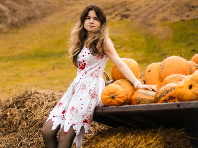 TinaRedly sits on a hay bale, wearing a white dress with red blood splatters, black tights, and long blonde hair. She's surrounded by large pumpkins on a wagon.