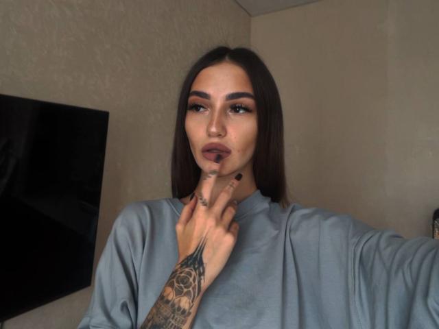SophieKitty heeft lang bruin haar, donkere wenkbrauwen en draagt een grijze trui. Ze raakt haar kin aan met een getatoeëerde vinger. Ze heeft volle lippen en donkere make-up..