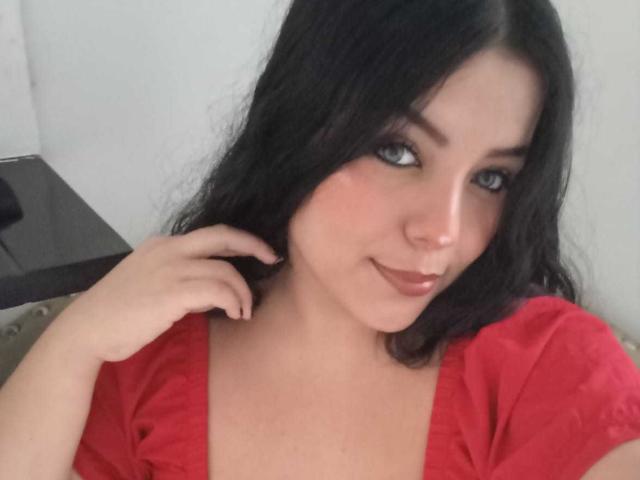 KonyBella has black hair, blue eyes, and a slight smile. She's wearing a red shirt, with her hand near her ear, looking at the camera. Her tits are visible.