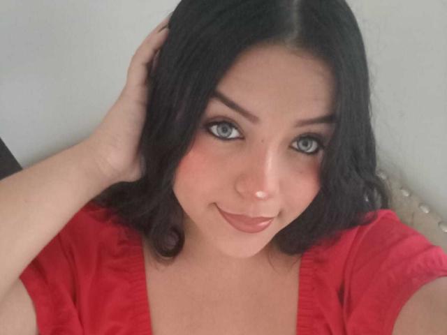 KonyBella has dark wavy hair, blue eyes, and a cute smile. She's wearing a red top, with her hand in her hair, looking directly at the camera.