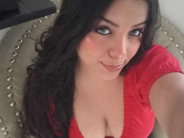 KonyBella has curly black hair, blue eyes, and a cute smile. She's wearing a red top with a low neckline, showing her big, round, sexy tits.