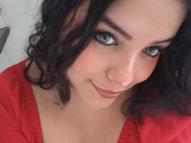 KonyBella with curly black hair, blue eyes, and a red shirt, smiling at the camera, showing a cute nose and full lips. Boobs slightly visible in the red shirt.