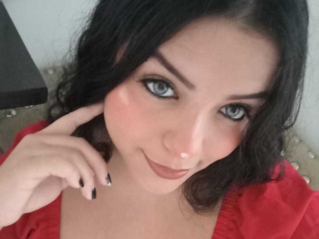 KonyBella has blue eyes, black wavy hair, and a cute smile. She's wearing a red top, with black nails touching her cheek. She's looking at the camera.