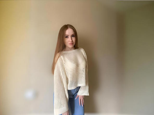 Emilysoo ha lunghi capelli castani, indossa una maglia bianca a maniche lunghe, lavorata all'uncinetto, che mette in mostra piccoli seni, e jeans blu, ed è in piedi con un leggero sorriso, in una stanza con una luce soffusa..