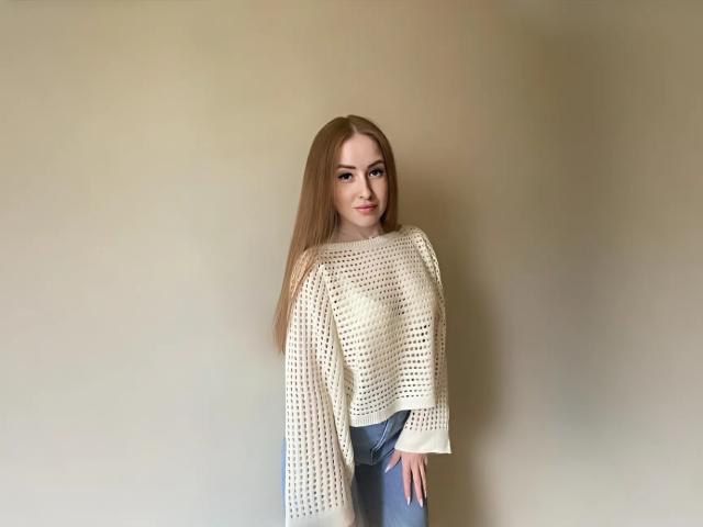 Emilysoo with long brown hair, wearing a white crochet top and blue jeans, standing against a plain wall, looking at the camera with a slight smile.