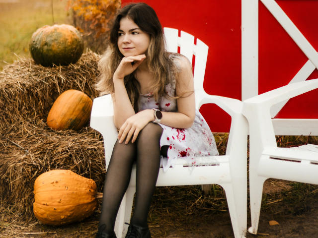 TinaRedly sits on a white plastic chair, leaning on her right hand with her chin resting on it. She has wavy, dark blonde hair and wears a white floral dress with black tights and black ankle boots. She looks to the right with a slight smile. Two large pumpkins are on the hay bale to her left. The background includes a red and white structure. She has a black wristwatch on her left wrist. The image has a natural, outdoor setting.