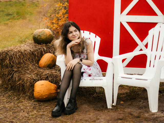 TinaRedly, with medium tits, sits on a white plastic chair, wearing a floral dress, black stockings, and black boots, in front of a red barn, with pumpkins and hay.
