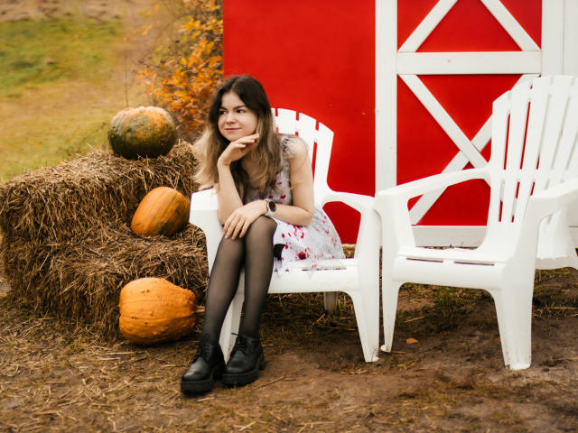 TinaRedly, com longos cabelos castanhos, vestido floral, botas pretas e collant pretos, sentada em uma cadeira de plástico branca, perto de um celeiro vermelho, fardos de feno e abóboras. Outono, relaxada, fofa..