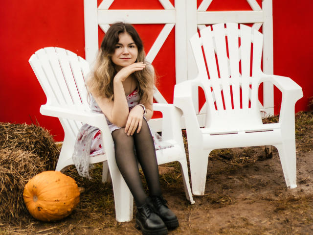 TinaRedly with wavy blonde hair, wearing a floral dress, black tights, and black shoes, sits on a white plastic chair, hand on chin, next to a pumpkin.