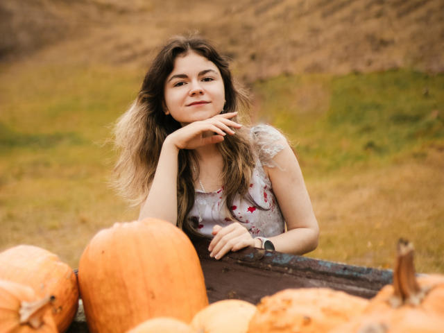 TinaRedly with long brown hair, white floral dress, resting chin on hand, smiling, surrounded by orange pumpkins, standing in front of rusty metal tray, looking cute and relaxed.