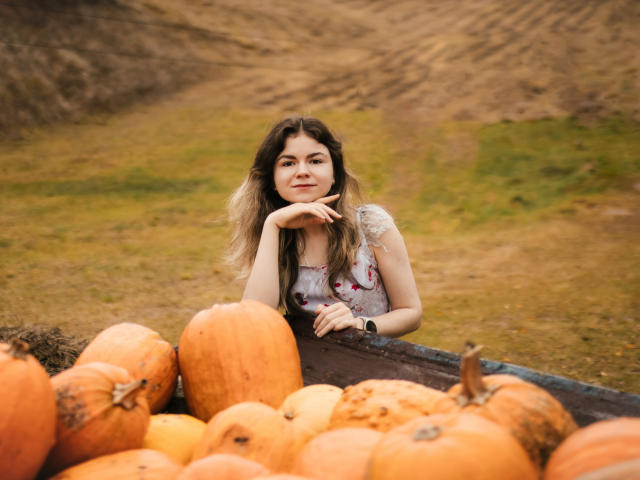 TinaRedly with puffy tits, hand on chin, leaning on pumpkin-filled wagon, wearing floral dress, brown wavy hair, smiling, wristwatch, autumn farm setting, orange pumpkins in foreground.