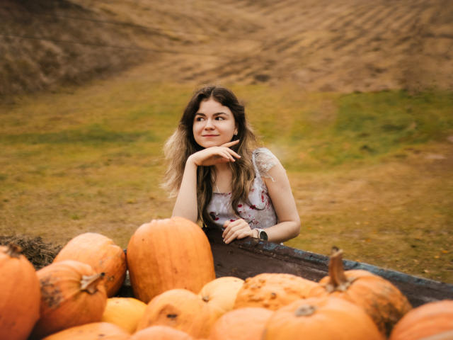 TinaRedly with wavy brown hair, wearing a floral dress, leaning on a pumpkin-filled wooden trough, smiling thoughtfully, hand on chin, autumn field background, orange pumpkins in foreground.