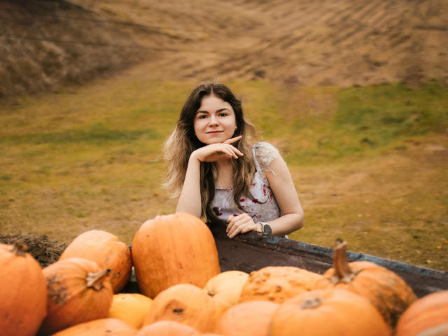 TinaRedly with long brown hair, wearing a floral dress, leans on a wagon filled with large orange pumpkins, smiling with her chin on her hand.