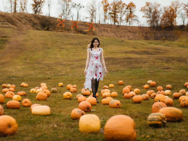 TinaRedly stands in a grassy field scattered with orange pumpkins. She wears a white floral dress with red patterns, black tights, and black shoes. Her dark hair is loose, and she looks slightly to the right with a neutral expression. The field has a slight hill in the background with orange crosses on a brown fence. The sky is overcast. The pumpkins are varied in size and placement, creating a scattered pattern. The image uses natural light and has a soft, autumnal color palette.