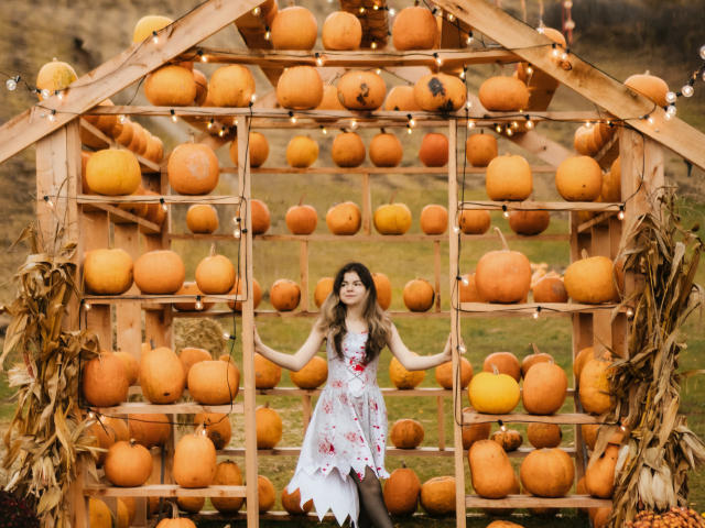 TinaRedly, avec ses longs cheveux bruns ondulés, portant une robe blanche à motifs floraux, se tient devant un cadre en forme de citrouille en bois, qu'elle tient, entourée de citrouilles oranges et de guirlandes lumineuses..