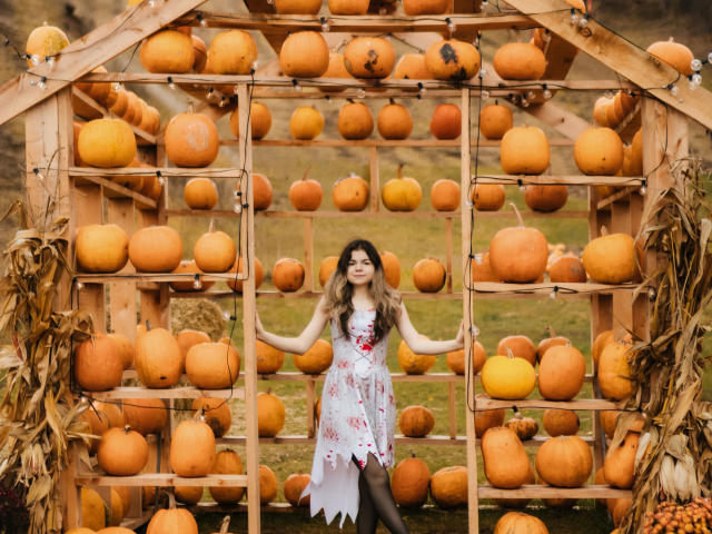 TinaRedly stands center in a wooden pumpkin rack, wearing a white floral dress with red accents, black leggings, and holding the rack's frame. She's surrounded by dozens of orange pumpkins. Her wavy brown hair is loose, and she smiles. The wooden rack is adorned with dried corn leaves. The composition is balanced with the pumpkins filling the frame, creating a rustic, autumnal vibe. Bright, natural light highlights the pumpkins and TinaRedly's dress.
