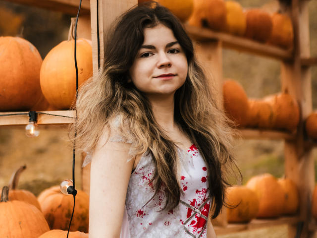 TinaRedly with long, wavy brown hair, wearing a white dress with red floral patterns, standing in front of wooden shelves filled with orange pumpkins, smiling softly.