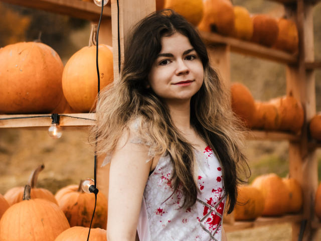 TinaRedly with long, wavy brown hair, wearing a white dress with red blood splatter, standing in front of a wooden shelf filled with pumpkins, smiling slightly.