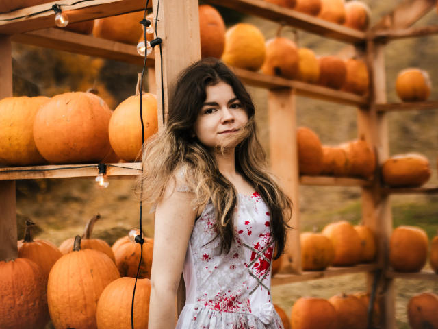 TinaRedly with long, wavy brown hair in a floral dress, standing next to wooden shelves filled with pumpkins, string lights above, and a calm, slightly dirty expression.