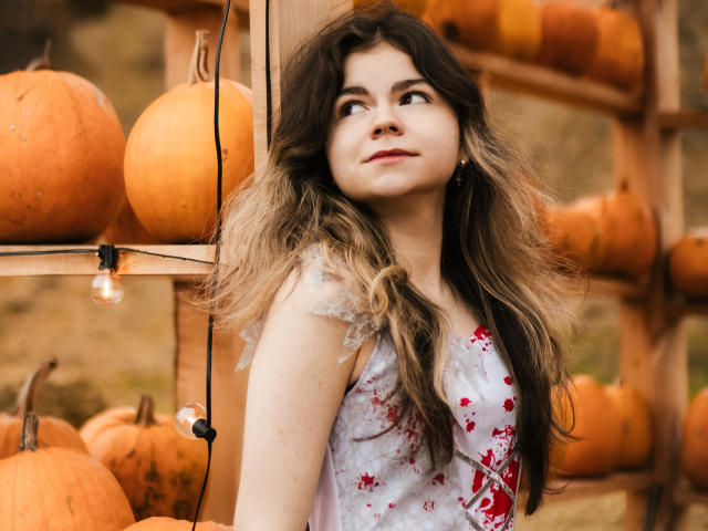 TinaRedly with long, wavy brown hair and light brown highlights, wearing a white dress with red floral patterns, looks to the right with a slight smile. She has light skin and is adorned with small earrings. Several orange pumpkins are stacked on wooden shelves behind her, with a single exposed light bulb hanging. The image has a warm, autumnal tone. The composition emphasizes TinaRedly's relaxed, confident posture and the vibrant pumpkins.