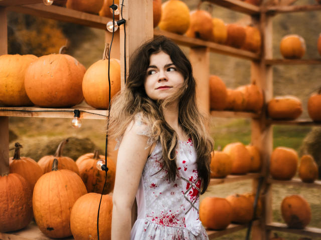 TinaRedly stands in a pumpkin display, wearing a white dress with red blood splatters. She has long, wavy brown hair with blonde highlights. Her expression is neutral, eyes looking slightly to the side. The background features wooden shelves filled with orange pumpkins. String lights hang from the shelves. The image is sharp, with natural light and a slightly warm, autumnal tone.