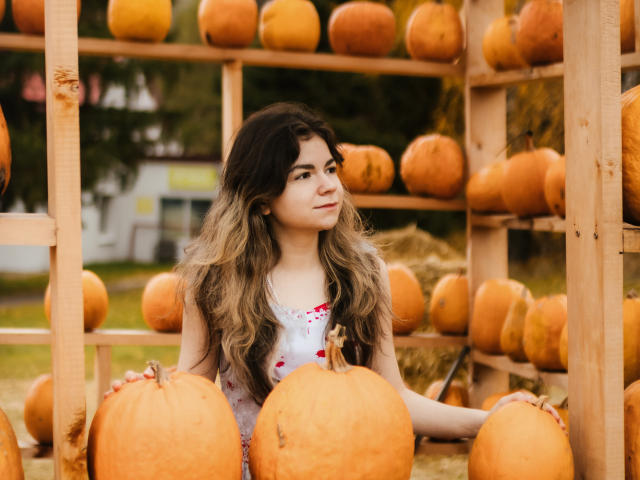 TinaRedly with long, wavy brown hair, wearing a white dress with red floral patterns, sitting among large orange pumpkins, looking to the side, surrounded by wooden shelves.