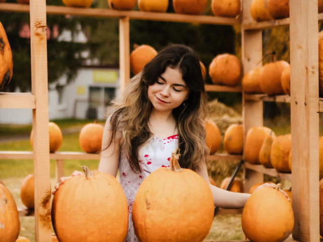 TinaRedly with long, wavy brown hair, wearing a white dress with pink flowers, smiling, holding a large orange pumpkin, surrounded by wooden shelves filled with more pumpkins.