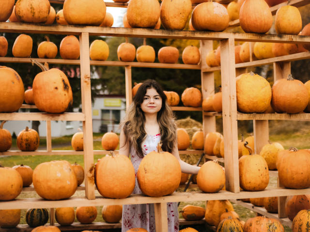 TinaRedly with long wavy hair, wearing a floral dress, stands among large orange pumpkins on wooden shelves, holding one with her right hand, smiling.