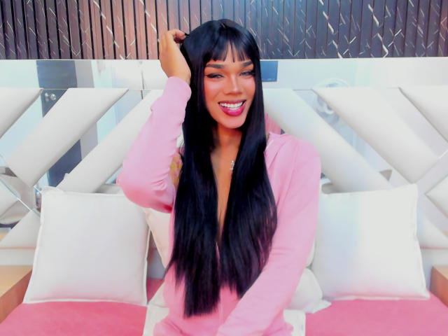 NaomiSteward with long black hair, pink long-sleeve shirt, smiling, hand in hair, white cushioned bed, pink and white pillows, wooden wall background, looking cute and sexy.