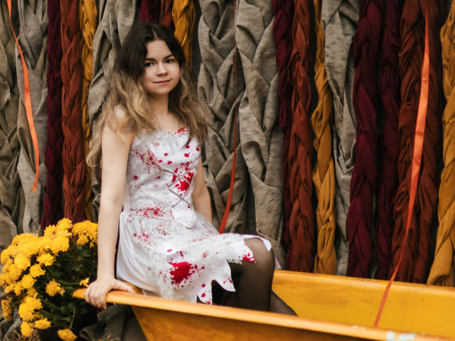 TinaRedly sits in a yellow boat, wearing a white floral dress with red stains. She has long, wavy, brown-to-blonde ombre hair and a slight smile. Her right hand holds a bouquet of yellow flowers. The background features vertically arranged, textured, colorful ribbons in gray, red, and orange. Her black tights are visible. The image has a natural, outdoor feel. Her relaxed posture and gentle expression create a calm, yet slightly mysterious, atmosphere.