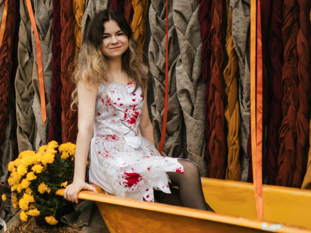 TinaRedly sits in a yellow boat, wearing a white dress with red blood-like stains. She has wavy, brown-to-blond ombre hair and a subtle smile. Black stockings contrast with her dress. Yellow flowers are in the boat's left side. The background has bunched, textured, multi-colored fabric. Orange ribbons hang from above. The composition is vibrant, with TinaRedly's dress and the flowers adding bright, contrasting colors. The textured fabric adds depth. The image is sharp, with clear, distinct elements.