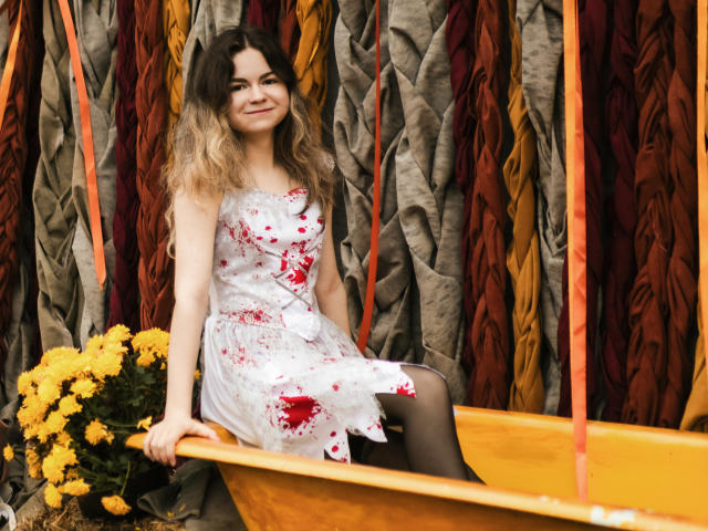 TinaRedly con un vestido blanco manchado de sangre, sentada en un bote amarillo, sosteniendo flores amarillas, sonriendo, cabello rubio ondulado, medias negras, fondo a rayas naranja y marrón..