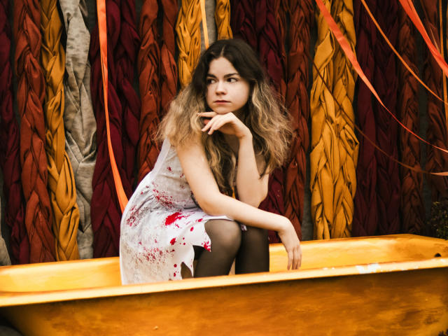Tina, mit welligem, braunem Haar, in einem weißen Kleid, das mit Rot gefleckt ist, schwarzen Strumpfhosen, sitzt in einer gelben Wanne, die Hand am Kinn, blickt zur Seite, vor einem farbenfrohen, blättrigen Hintergrund..