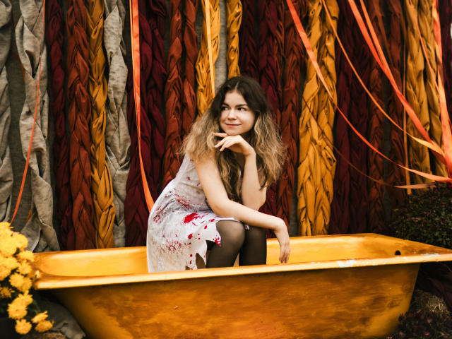 A photograph of TinaRedly, a young woman with wavy brown hair, sitting in a yellow, metallic basin. She's wearing a white dress with red floral patterns, black tights, and has a contemplative expression. Her right hand rests on her chin while her left arm is on the basin's edge. The background features vertically hung, ruched fabric in red, orange, and yellow, with yellow flowers in the lower left corner. The image has a warm, natural light and a casual, relaxed atmosphere. The fabric's texture contrasts with the smooth basin. TinaRedly's dress has a slight sheen, and the composition is balanced.