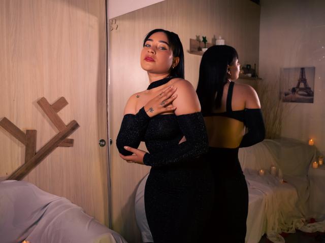 SaharaArisa stands in a black, off-shoulder, glitter dress, covering her tits with one hand, her ass slightly visible, reflection in mirror, wooden wall, candles in background.