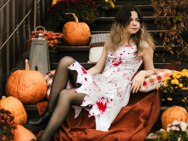 TinaRedly sits on a red cloth, surrounded by pumpkins and autumn flowers. She wears a white dress with red blood splatters, black tights, and black boots. Her wavy blonde hair is loose. The stairs are dark, and a gray lantern is to her left. The composition uses warm, autumnal colors, and TinaRedly's relaxed, slightly bored expression adds a contrast. The image has a realistic, slightly gritty style.