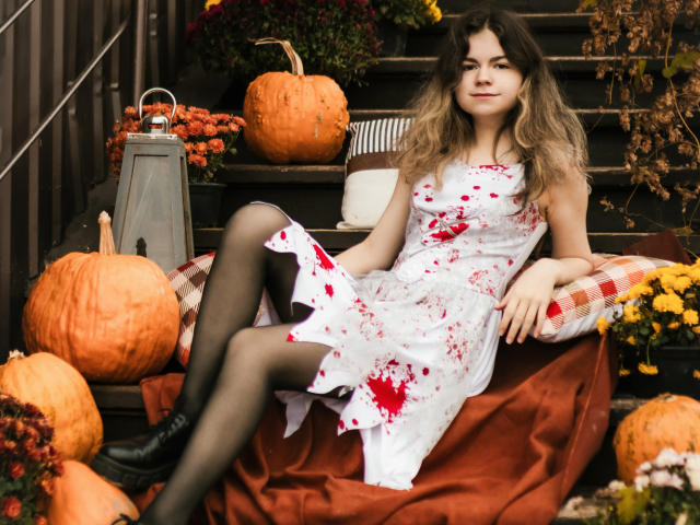 TinaRedly is a young woman with long, wavy brown hair sitting on a red cloth. She wears a white dress splattered with red blood-like spots, black stockings, and black boots. She's surrounded by pumpkins, a lantern, and fall flowers. She's leaning back with a relaxed, confident expression. The image uses a warm, autumnal color palette and has a slightly messy, rustic feel. Her casual, confident posture contrasts with the spooky, seasonal elements.