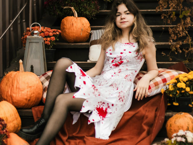 TinaRedly, a young woman with wavy brown hair, sits on a set of dark wooden steps. She wears a white dress with red blood splatters, black pantyhose, and black boots. She's relaxed, leaning back on a red and white checkered pillow. Surrounding her are several pumpkins, a lantern, and autumnal flowers. The composition uses warm, autumnal colors and contrasts TinaRedly's white dress with the red splatters and her black boots. The image has a casual, slightly rustic vibe.