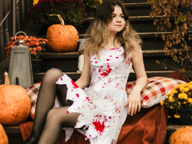 TinaRedly sits on a set of dark wooden steps, wearing a white dress splattered with red blood-like spots and black tights. Her wavy brown hair is loose, and she looks to the right with a slight smile. Pumpkins and yellow flowers are on the steps and a lantern is on the left. A red and white checkered pillow is under her. The image has a realistic, slightly rustic style.