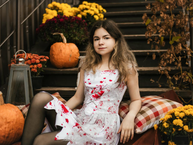 TinaRedly, with wavy brown hair, wears a bloody white floral dress and black tights, sitting on a plaid pillow, surrounded by pumpkins and yellow flowers, looking at camera.