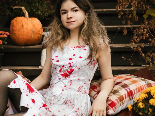 TinaRedly sits on outdoor stairs, wearing a white dress with red floral patterns. Her wavy, blonde-brown hair falls around her face. A small white button decorates the dress. She's on a plaid pillow with orange and brown stripes. A pumpkin and dried plants are in the background. Her right arm rests on a yellow flower. The image has a natural, autumnal feel. She looks calm and slightly smiling. Her skin is fair, and she has light makeup. The photograph is realistic.