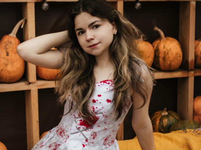 TinaRedly, a young woman with long, wavy brown hair and fair skin, sits in a wooden shelf filled with pumpkins. She wears a white dress with red blood splatters, looking directly at the camera with a slight smile. Her right arm is raised, hand resting on the back of her head. The background has an orange-yellow cloth on the lower right. The image uses natural lighting. Her relaxed, confident pose contrasts with the eerie, Halloween-themed attire.