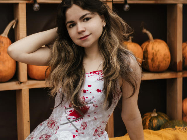 TinaRedly has long, wavy brown hair with blonde highlights. She's wearing a white dress with red floral patterns, sitting with one hand in her hair. Her expression is relaxed, slightly smiling. The background has wooden shelves with large, orange pumpkins. The colors are warm, with a mix of orange, brown, and white. TinaRedly's skin is light, and she has small earrings. The image has a natural, slightly candid feel.