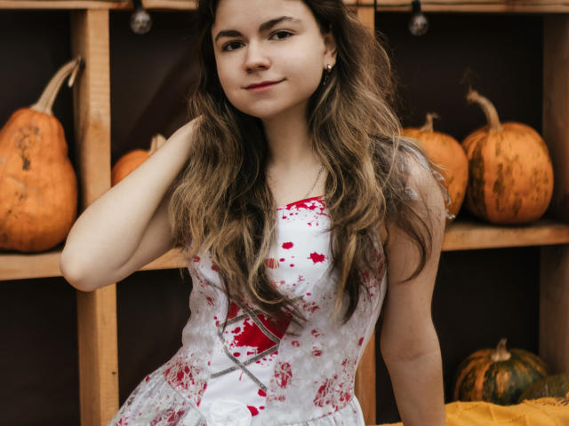 TinaRedly stands in front of wooden shelves with pumpkins. She has long, wavy brown hair and fair skin. She wears a white dress with red blood splatter patterns and a lace-up detail. Her right arm is raised, touching her hair. She has a slight, confident smile. The dress contrasts with her skin tone, and the blood splatter adds a dark, dramatic element. The wooden shelves and pumpkins create a rustic, autumnal setting. The image is sharply focused with natural lighting.