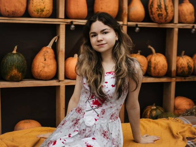 TinaRedly with long wavy brown hair, wearing a white floral dress with red splashes, sitting on a yellow blanket, surrounded by various pumpkins in a wooden shelf.