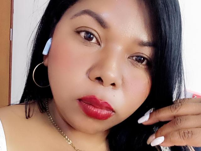 Keilathompson has a cute face, red lipstick, black hair, hoop earrings, white earbuds, and a gold necklace. She's looking at the camera with a slight pout.