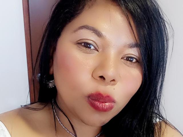 Keilathompson's face, showing dark wet hair, shiny skin, red lipstick, small nose, and brown eyes, with a slight pout and a silver necklace. She has black earrings and a white top.